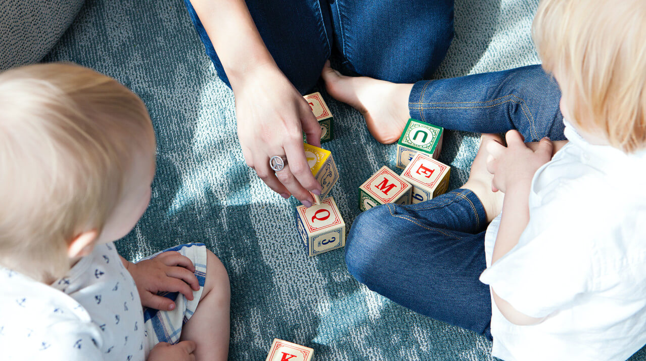 Erfahrungsbericht zu Pflegekinder: “Ich kann mir nicht vorstellen, dass ich ein leibliches Kind mehr lieben könnte.” Zwei kleine Kinder spielen mit einer Erwachsenen. Symbolbild für Erfahrungsbericht einer Pflegefamilie